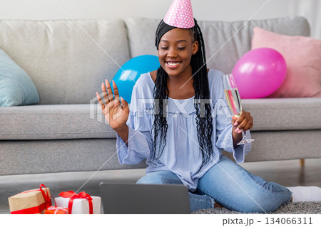 Happy african american young lady with glass of champagne and festive hat having online party from home, using laptop, sitting on floor next to giftboxes and colorful balloons, copy space Happy african american young lady with glass of champagne and festive hat having online party from home, using laptop, sitting on floor next to giftboxes and colorful balloons, copy space 134066311