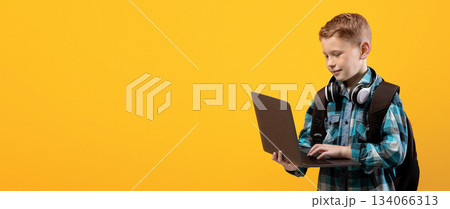 Technologies in school education. Smart schoolboy with backpack and headset holding laptop over yellow studio background, copy space. Cute kid using laptop for studying, doing homework, websurfing Technologies in school education. Smart schoolboy with backpack and headset holding laptop over yellow studio background, copy space. Cute kid using laptop for studying, doing homework, websurfing 134066313