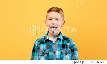 Spoiled ginger kid cute school boy in casual outfit showing his tongue at camera over yellow studio background, copy space. Spoiled child, kids behaviour problems concept 134066324