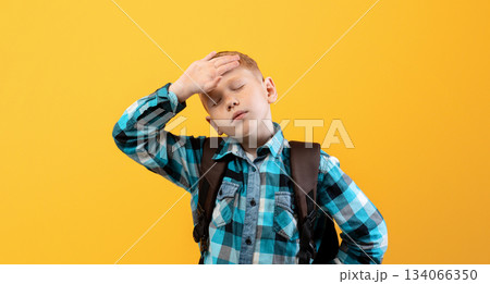 Exhausted kid schooler with heavy backpack touching his forehead on yellow studio background. Tired redhead boy in plaids shirt got tired of studying, suffering from burnout in school 134066350