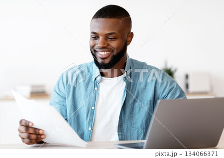 Millennial Black Freelancer Man Working With Papers At Desk In Home Office. Smiling African American Guy Sitting At Table With Computer Reading Documents And Checking Financial Reports, Free Space Millennial Black Freelancer Man Working With Papers At Desk In Home Office. Smiling African American Guy Sitting At Table With Computer Reading Documents And Checking Financial Reports, Free Space 134066371