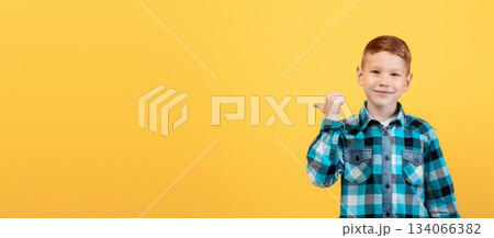 Smiling red-haired caucasian boy aiming at free space behind his back, empty space for text or advertisement, panorama. Cheerful ginger kid pointing with thumb at copy space over yellow background Smiling red-haired caucasian boy aiming at free space behind his back, empty space for text or advertisement, panorama. Cheerful ginger kid pointing with thumb at copy space over yellow background 134066382