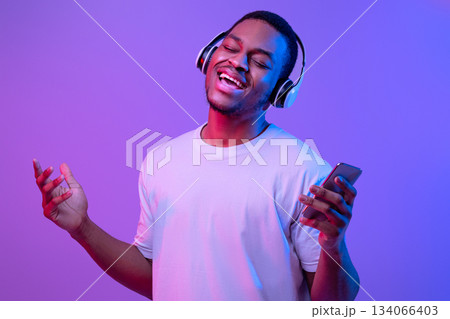 Joyful black guy in wireless headphones listening music online on smartphone and singing under futuristic neon lighting, enjoying favorite songs, standing with closed eyes on fluorescent background Joyful black guy in wireless headphones listening music online on smartphone and singing under futuristic neon lighting, enjoying favorite songs, standing with closed eyes on fluorescent background 134066403