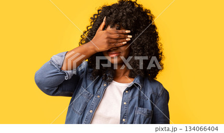 Curly black woman covering eyes with one hand, feeling scared or anxious, wondering or blindly waiting for a surprise against yellow studio background, panorama with copy space 134066404
