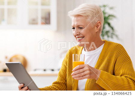 Joyful old grey-haired woman eating healthy food and using digital tablet in kitchen, empty space. Happy grandmother with glass of fresh juice reading culinary blog on modern pad Joyful old grey-haired woman eating healthy food and using digital tablet in kitchen, empty space. Happy grandmother with glass of fresh juice reading culinary blog on modern pad 134066425