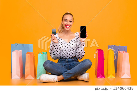 Online Shopping Concept. Excited overjoyed lady showing smartphone and credit card to camera, ordering clothes and purchases via internet, sitting with colorful paper bags at pastel orange studio Online Shopping Concept. Excited overjoyed lady showing smartphone and credit card to camera, ordering clothes and purchases via internet, sitting with colorful paper bags at pastel orange studio 134066439