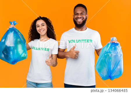 Couple Of Multiethnic Volunteers Holding Blue Plastic Garbage Bags Picking Litter Gesturing Like Standing Over Yellow Studio Background. Volunteering Work, Let's Keep Planet Environment Clean Concept Couple Of Multiethnic Volunteers Holding Blue Plastic Garbage Bags Picking Litter Gesturing Like Standing Over Yellow Studio Background. Volunteering Work, Let's Keep Planet Environment Clean Concept 134066485