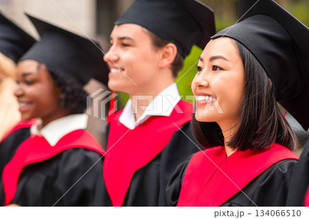 Multiracial group of cheerfully graduates having graduation ceremony, wearing black and red graduation robes and caps, standing in a row at university campus, closeup, panorama 134066510