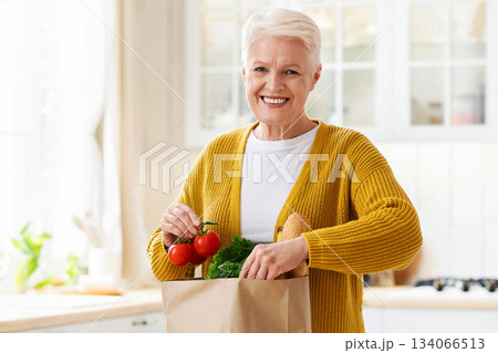 Happy senior woman checking paper bag with grocery ordered from internet during COVID-19 pandemic, kitchen interior, copy space. Smiling old lady unpacking purchase after shopping. Food delivery Happy senior woman checking paper bag with grocery ordered from internet during COVID-19 pandemic, kitchen interior, copy space. Smiling old lady unpacking purchase after shopping. Food delivery 134066513