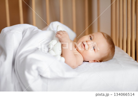 Good morning world. Happy baby boy waking up after sleeping in wooden crib, laying under blanket and smiling, holding white toy, side view. Cute baby girl laying in toddler bed 134066514
