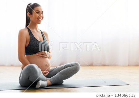 Young expecting woman training on mat at home, getting ready for childbirth. Smiling beautiful pregnant female exercising in living room, tenderly embracing tummy and looking away, copy space 134066515