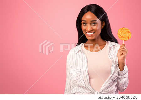 Portrait of beautiful African American lady holding lollipop, looking at camera and smiling on pink studio background. Young black woman with candy on a stick having sweet tooth 134066528