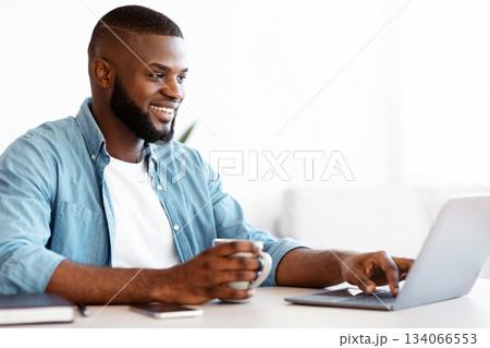 Freelancing Concept. Cheerful Black Freelancer Man Drinking Coffee And Working On Laptop At Home Office, Millennial African American Guy Sitting At Desk With Computer, Enjoying Remote Job, Copy Space Freelancing Concept. Cheerful Black Freelancer Man Drinking Coffee And Working On Laptop At Home Office, Millennial African American Guy Sitting At Desk With Computer, Enjoying Remote Job, Copy Space 134066553