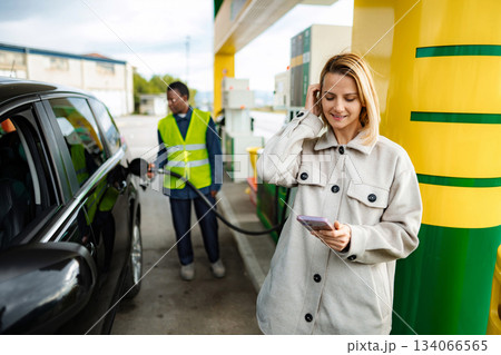 Woman using smartphone while gas station attendant refueling car 134066565