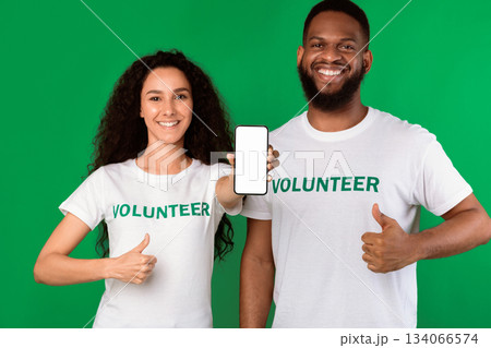 Two Cheerful Volunteers Showing Phone Empty Screen Gesturing Like Approving Charity Application Standing On Green Studio Background, Smiling To Camera. Volunteering Work, Call Us Concept. Mockup Two Cheerful Volunteers Showing Phone Empty Screen Gesturing Like Approving Charity Application Standing On Green Studio Background, Smiling To Camera. Volunteering Work, Call Us Concept. Mockup 134066574