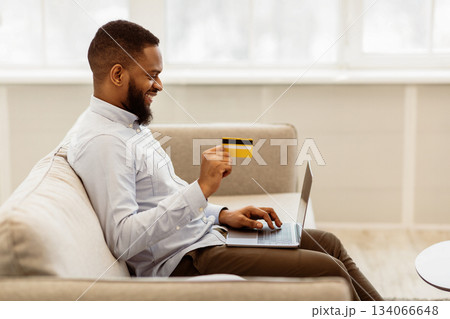 Easy Payment And Cashback Concept. Side view portrait of smiling african american young man holding credit card in hand and using laptop, sitting on couch at home, typing on keyboard, copy space Easy Payment And Cashback Concept. Side view portrait of smiling african american young man holding credit card in hand and using laptop, sitting on couch at home, typing on keyboard, copy space 134066648