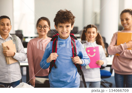 Return Back To School. Smiling small boy wearing backpack posing in classroom, diverse group of happy international classmates standing in line in the blurred background, studying and learning 134066709