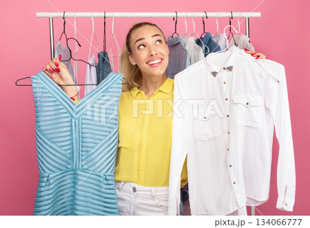 Dilemma Concept. Portrait of young pensive lady choosing trendy outfit, holding hangers with dress and shirt, thinking about purchase, making decision what to wear, rack in the background 134066777