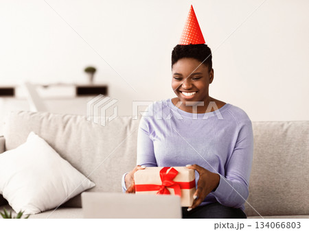 Virtual Party. Positive Cheerful African American Woman Wearing Hat, Showing Wrapped Gift Box With Present To Computer Webcam While Having Online Video Conference, Congratulating Friend With Holiday 134066803