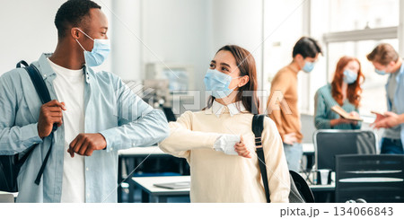 Elbow Bump. Smiling multicultural students wearing protective face masks and backpacks greeting each other. Woman and african american guy avoid touching, studying in university, standing at classroom Elbow Bump. Smiling multicultural students wearing protective face masks and backpacks greeting each other. Woman and african american guy avoid touching, studying in university, standing at classroom 134066843