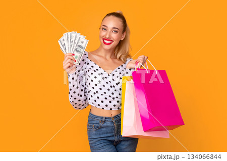 Commerce Concept. Portrait of smiling young lady holding bunch of cash money fan and shopping bags in hand, looking at camera, standing at studio, isolated over yellow background. Cashback 134066844