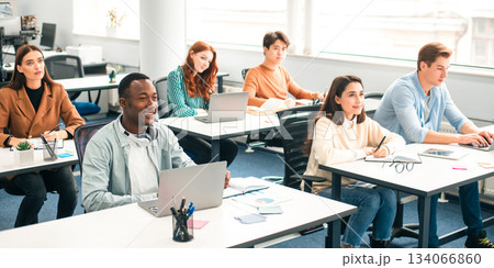 Lecture Concept. Group of interested multicultural mixed race group of students sitting at tables with pc in modern classroom, listening to teacher, taking notes writing in notebooks, back to study Lecture Concept. Group of interested multicultural mixed race group of students sitting at tables with pc in modern classroom, listening to teacher, taking notes writing in notebooks, back to study 134066860