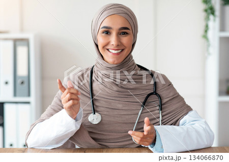 Smiling female doctor young beautiful woman in hijab with stethoscope making consultation in her cabinet, sitting at workdesk with medical chart, notebook and laptop in clinic, copy space 134066870