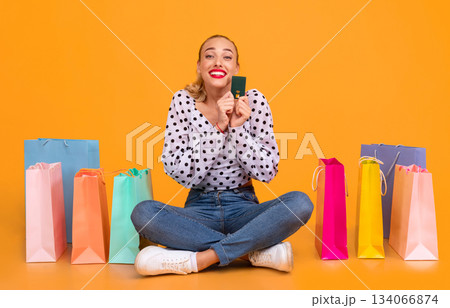 Easy Payment. Portrait of excited stylish young woman holding credit card, ordering purchases and paying online via internet, buying clothes, sitting at yellow studio on the floor with shopping bags Easy Payment. Portrait of excited stylish young woman holding credit card, ordering purchases and paying online via internet, buying clothes, sitting at yellow studio on the floor with shopping bags 134066874