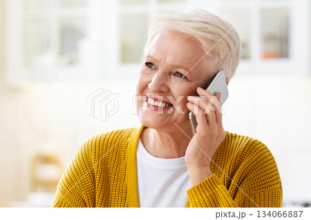 Happy attractive elderly woman having healthy lunch and talking on mobile phone, white kitchen interior. Smiling old lady having conversation on smartphone during lunch time, looking at copy space 134066887