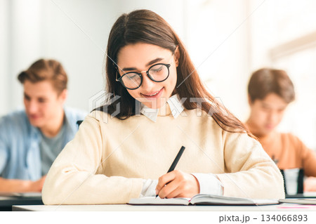 Studying And Learning Concept. Portrait of smiling young woman in eyeglasses sitting at table in lecture hall in highschool, writing in notebook, taking notes or examination, selective focus Studying And Learning Concept. Portrait of smiling young woman in eyeglasses sitting at table in lecture hall in highschool, writing in notebook, taking notes or examination, selective focus 134066893
