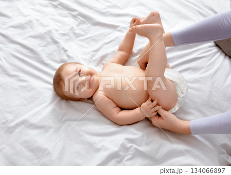Unrecognizable mother changing her smiling little baby's diaper on white, top view. Cute toddler blonde boy having fun while his mom dressing him up, playing with his feet, closeup, empty space. 134066897