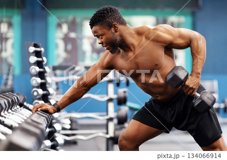 Motivated african american shirtless bodybuilder training arms and back muscles with dumbbells at modern gym, empty space. Hot black young man working on his biceps, lifting barbell with one hand 134066914