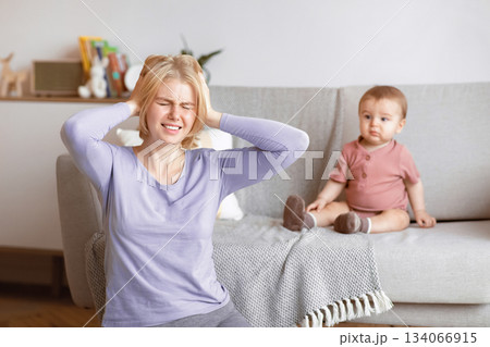 Postnatal depression concept. Stressed blonde woman young mother suffering from headache, sitting with baby at home, touching her head and screaming. Motherhood, multi-tasking and family Postnatal depression concept. Stressed blonde woman young mother suffering from headache, sitting with baby at home, touching her head and screaming. Motherhood, multi-tasking and family 134066915