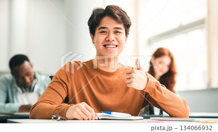 Education Concept. Portrait of smiling asian male student sitting at desk in classroom at university, showing thumbs up sign gesture, looking posing at camera, selective focus. Academic Success Education Concept. Portrait of smiling asian male student sitting at desk in classroom at university, showing thumbs up sign gesture, looking posing at camera, selective focus. Academic Success 134066932