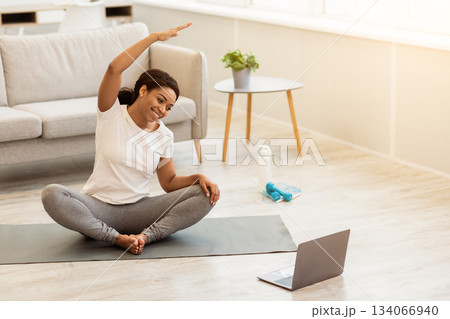 Stay Fit Concept. Smiling african american woman exercising at home, stretching arms, watching online training on laptop compute in living room on the floor on yoga mat, copy space Stay Fit Concept. Smiling african american woman exercising at home, stretching arms, watching online training on laptop compute in living room on the floor on yoga mat, copy space 134066940