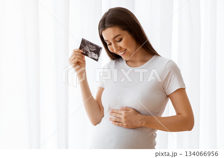 Maternity Concept. Happy Beautiful Pregnant Lady Holding Ultrasound Photo Of Her Baby And Tenderly Embracing Belly While Standing Near Window At Home, Expectant Mother Keeping Sonography Scan 134066956