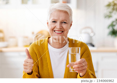Cheerful senior grey-haired woman with short haircut holding glass of water showing thumb up, enjoying healthy lifestyle while retirement, sitting at white kitchen at home, copy space Cheerful senior grey-haired woman with short haircut holding glass of water showing thumb up, enjoying healthy lifestyle while retirement, sitting at white kitchen at home, copy space 134066962
