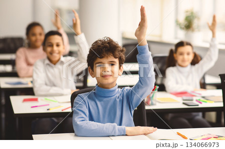 First Day Of School. Portrait of cute boy sitting at table in classroom, group of multicultural small pupils all raising hands up for an answer, studying in modern classroom, selective focus First Day Of School. Portrait of cute boy sitting at table in classroom, group of multicultural small pupils all raising hands up for an answer, studying in modern classroom, selective focus 134066973