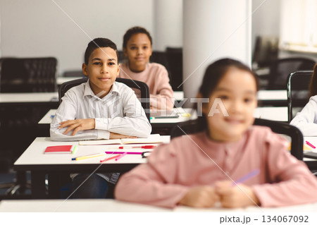 Elementary Education. Portrait of small smiling boy sitting at table in classroom at primary school or kindergarten with group of classmates. Return back to school after covid quarantine Elementary Education. Portrait of small smiling boy sitting at table in classroom at primary school or kindergarten with group of classmates. Return back to school after covid quarantine 134067092