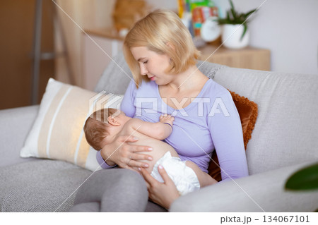 Beautiful blonde woman holding her little kid, sitting on sofa in living room, breastfeeding, copy space. Peaceful young mother nursing her adorable baby in diaper, home interior 134067101