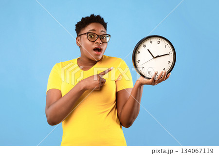 Deadline, Time Is Over. Oversized African Woman Showing Clock Posing Standing Over Blue Studio Background, Looking At Camera. Shocked Black Lady Being Late. Time's Tickling Concept 134067119