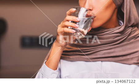 Muslim Woman Drinking Water From Glass Standing In Modern Kitchen At Home, Wearing Hijab Headscarf. Stay Hydrated. Healthy Lifestyle, Hydration And Daily Water Intake Concept Muslim Woman Drinking Water From Glass Standing In Modern Kitchen At Home, Wearing Hijab Headscarf. Stay Hydrated. Healthy Lifestyle, Hydration And Daily Water Intake Concept 134067122