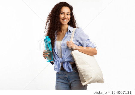 Happy Woman Carrying White Eco Bag Holding Reusable Bottle Of Water Posing Smiling To Camera Standing Over White Studio Background. Minimalism, Ecology Friendly Lifestyle. No More Plastic Concept Happy Woman Carrying White Eco Bag Holding Reusable Bottle Of Water Posing Smiling To Camera Standing Over White Studio Background. Minimalism, Ecology Friendly Lifestyle. No More Plastic Concept 134067131