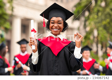 Cheerful african american young lady in graduation costume showing her diploma and smiling at camera, black female student posing over international group of students at university campus, copy space Cheerful african american young lady in graduation costume showing her diploma and smiling at camera, black female student posing over international group of students at university campus, copy space 134067142