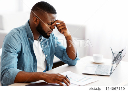 Migraine Concept. Overworked African American Man Suffering From Headache At Workplace, Stressed Black Freelancer Guy Sitting At Desk With Laptop And Documents And Touching Nose Bridge, Feeling Unwell 134067150