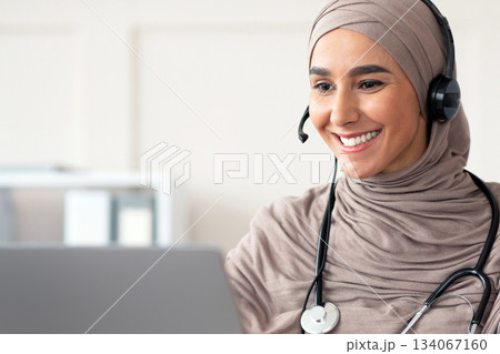 Cheerful muslim lady doctor having video chat with patient, using headset and laptop, talking to sick client online while working at clinic, copy space. Female physician having online appointment Cheerful muslim lady doctor having video chat with patient, using headset and laptop, talking to sick client online while working at clinic, copy space. Female physician having online appointment 134067160