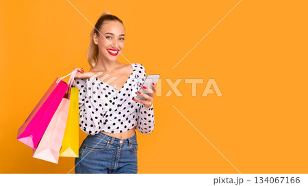 Fashion And Style. Smiling female model carrying colorful shopping bags and holding smartphone, making purchases in online store standing isolated over pastel orange studio background, copy space Fashion And Style. Smiling female model carrying colorful shopping bags and holding smartphone, making purchases in online store standing isolated over pastel orange studio background, copy space 134067166