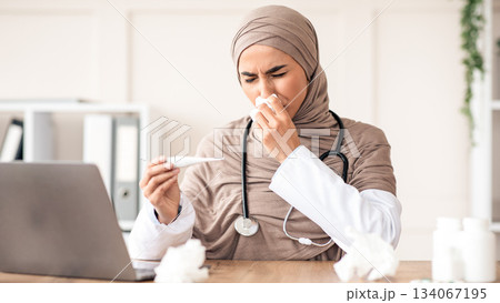 Ill doctor young muslim woman in hijab sneezing nose and checking fever while sitting at her workdesk in front of laptop, got sick during flu or COVID-19 pandemic, clinic interior 134067195