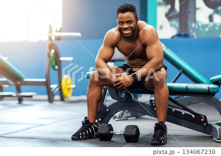 Cheerful shirtless black guy sitting on bench and smiling at camera, resting after biceps work out, gym interior, copy space. Happy african american sportsman enjoying exercises with barbells Cheerful shirtless black guy sitting on bench and smiling at camera, resting after biceps work out, gym interior, copy space. Happy african american sportsman enjoying exercises with barbells 134067230