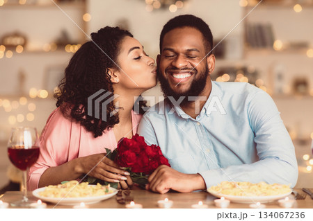 Celebrating Special Day. Beautiful black girlfriend kissing her smiling boyfriend in cheek, holding bouquet of red roses. Couple sitting at table in luxury restaurant during romantic date and dinner Celebrating Special Day. Beautiful black girlfriend kissing her smiling boyfriend in cheek, holding bouquet of red roses. Couple sitting at table in luxury restaurant during romantic date and dinner 134067236
